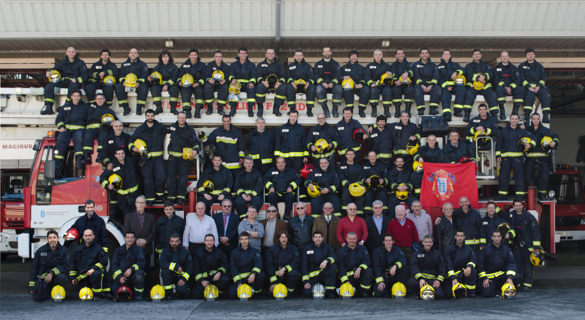 El cuerpo de bomberos al completo y pertrechados con sus trajes de intervención, en el lateral del camión escalera ocupando toda su estructura.