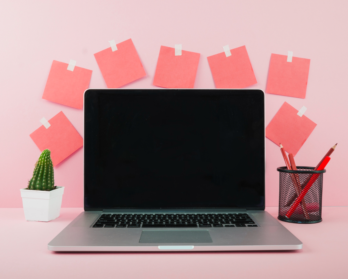  Ordenador portátil con pantalla negra sobre escritorio rosa. Con elementos decorativos ao carón como cactus, lapiceiro con lápis e possits 