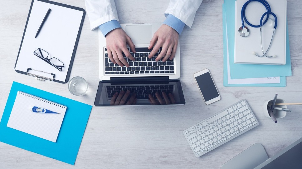 Persoa con bata de sanitario escribindo nun ordenador.