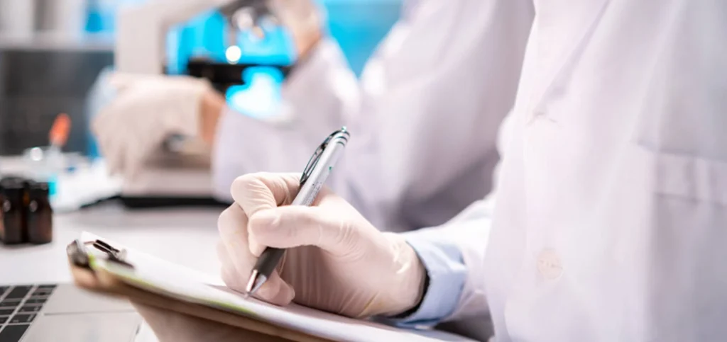 Persona anotando en una hoja datos en un laboratorio, lleva una bata y guantes de latex