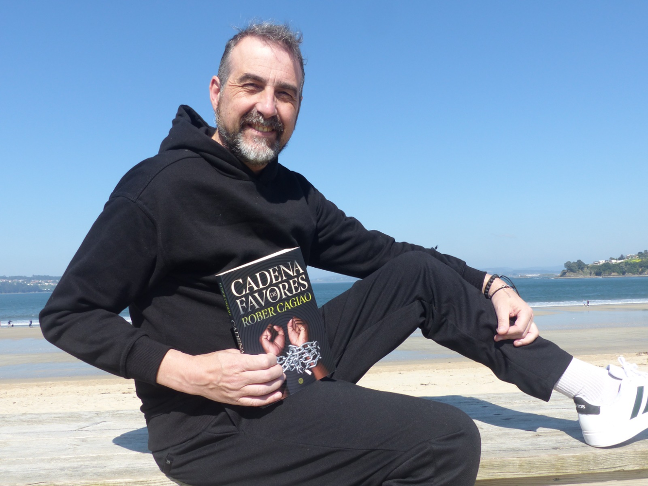 Fotografía de Rober Cagiao, vestido de negro e sentado diante dunha praia e cun libro seu na man dereita